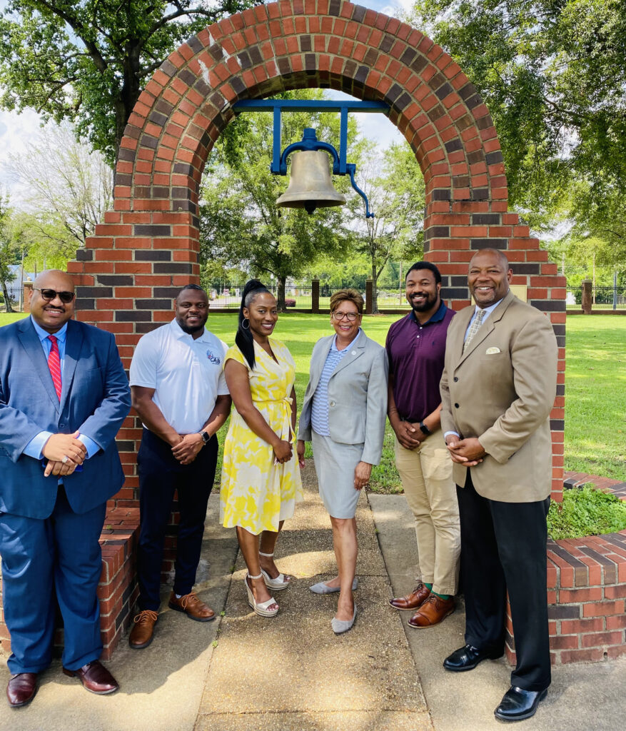 A group of black educators stand in front of a bell monument outdoors