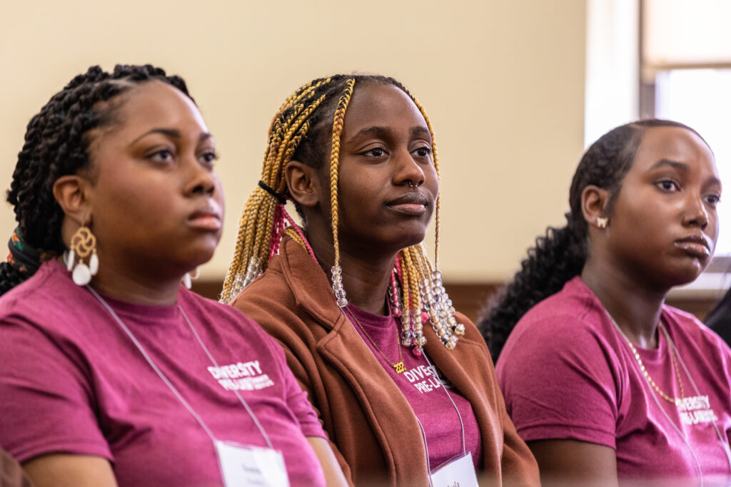 Three Black female college students sit in a courtroom and listen to a presentation.