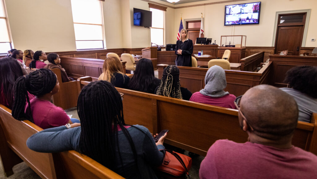 A group of Black female college students listen to a judge speaking in a courtroom