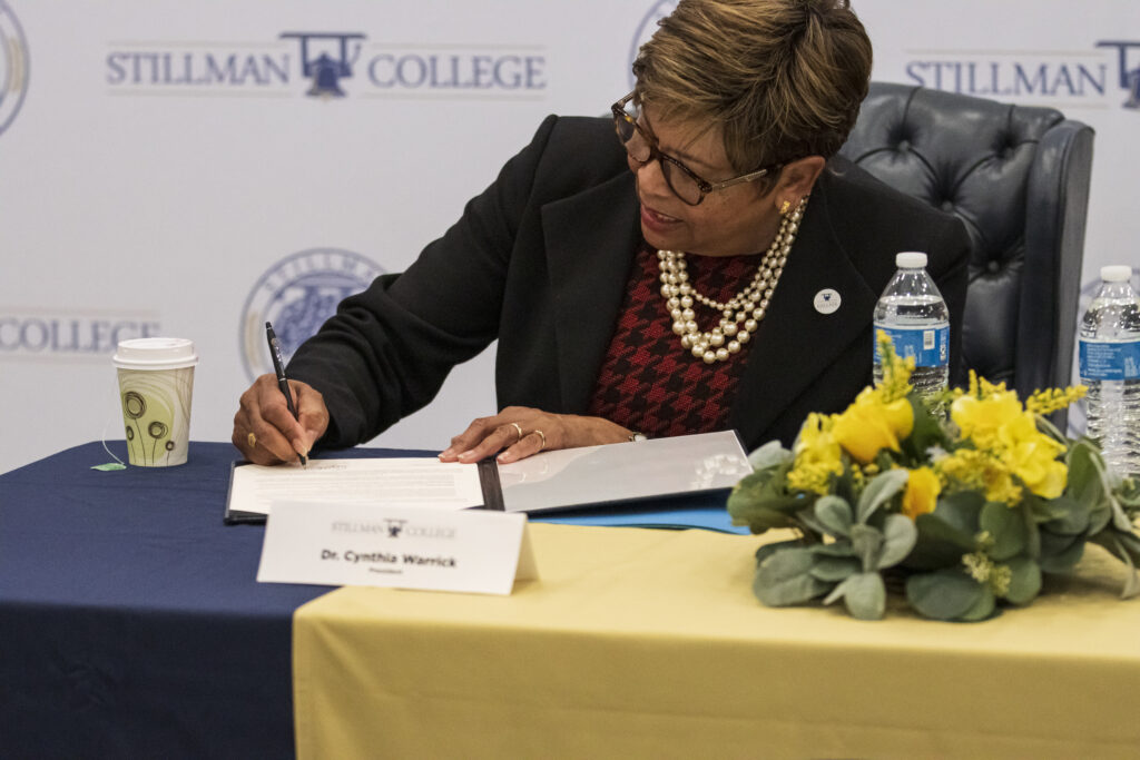 A black woman signs paperwork during a ceremony indoors.