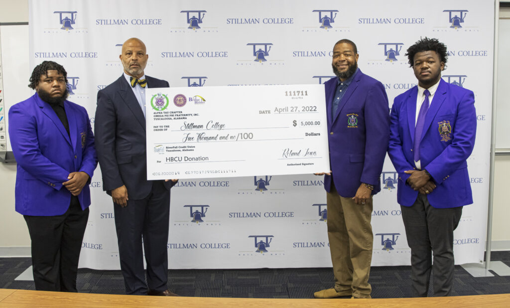 Four black men hold an oversized check and pose for a photo indoors