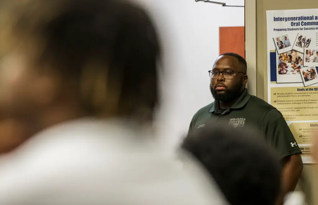 A Black man stands against a wall in a classroom and listens to a speech.