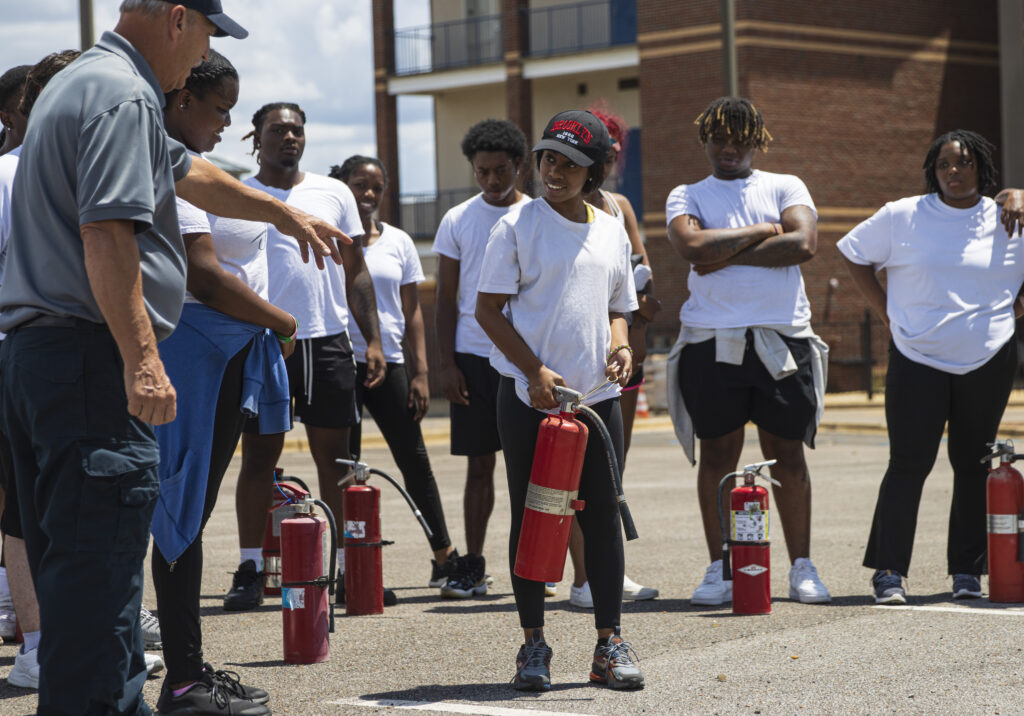 A Black female college student holds a fire extinguisher while listening to a firefighter speak