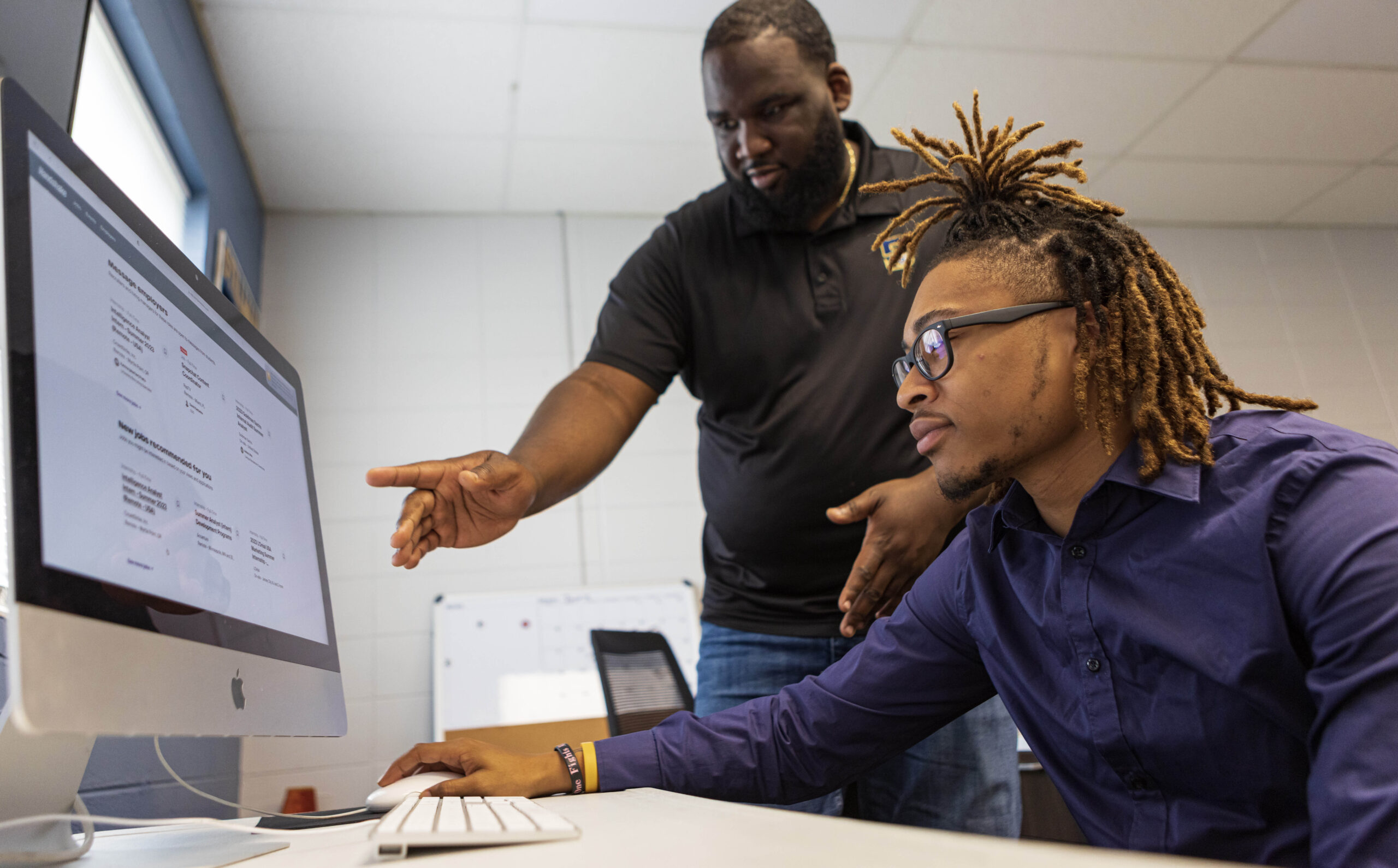 A Black man points to a computer screen as a young black male college student looks on.