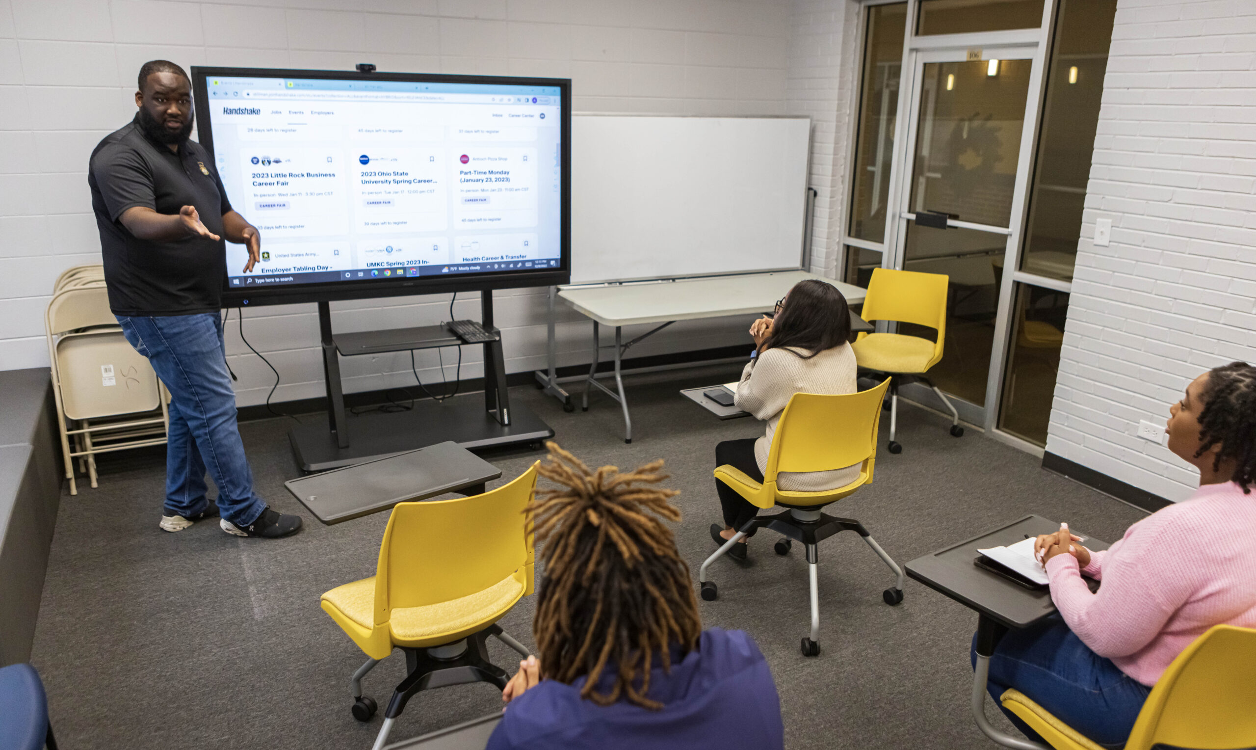 A black college instructor stands at an interactive board while teaching three black students