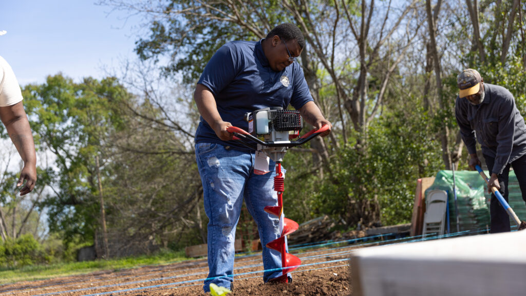 A Black male college student uses an auger to prep soil