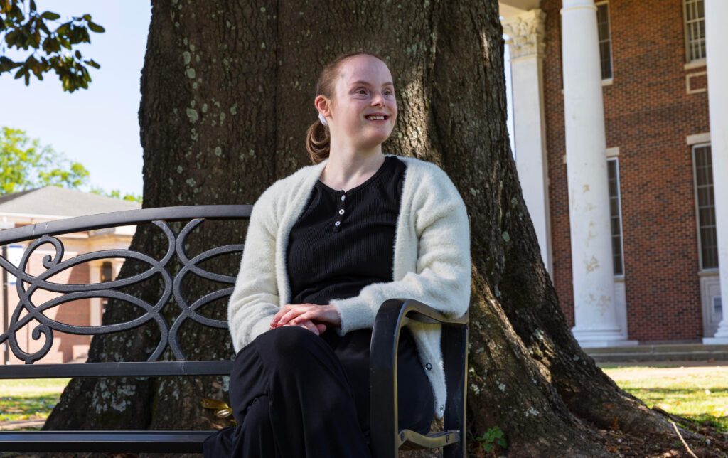 A white female college student poses for a picture while sitting on a bench under a tree