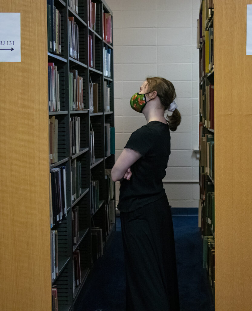 A white female college student browses books on a library shelf.