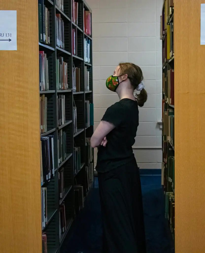 A white female college student browses books on a library shelf.