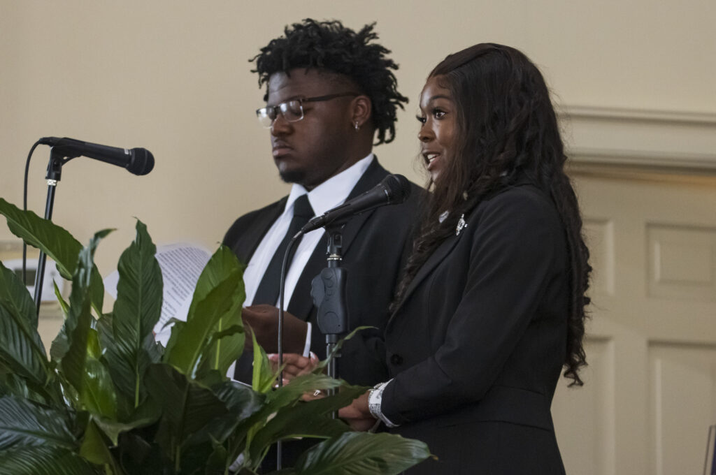 A black male college student and black female college student speak at a lectern at a church