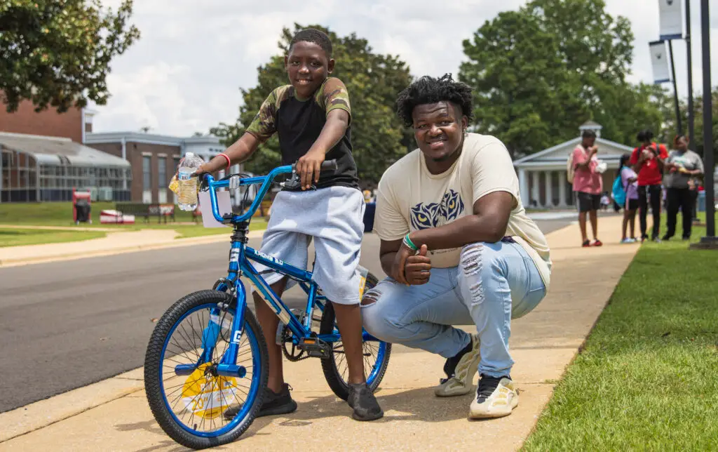 A black male college student poses for a picture next to a child on a bicycle